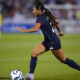 A woman in a blue jersey dribbles the ball on a soccer field